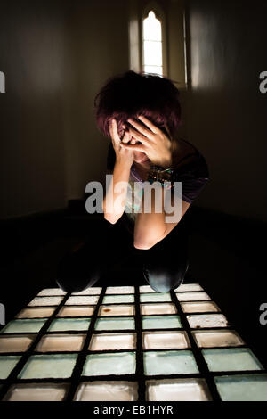 A despairing young teenage girl woman in a prison jail cell type location with light streaming through a barred window Stock Photo