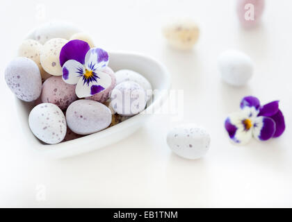 Pastel colourful mini eggs in a white heart shaped bowl with viola pansy on a white blurred backdrop Stock Photo