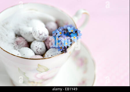 Pastel colourful mini eggs in a vintage teacup with white feather and forget-me-nots on a pink blurred backdrop Stock Photo