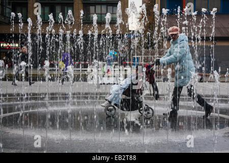 Paddington Basin, London, UK. November 25th 2014. UK Weather: A chilly start to the day in London. Pictured: A woman pushes a child in a buggy betwen the jets of a fountain in Paddington Basin. Credit:  Paul Davey/Alamy Live News Stock Photo