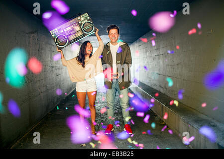 Caucasian couple carrying boom box and throwing confetti in tunnel Stock Photo