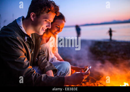 Caucasian couple using cell phone together near fire on beach Stock Photo