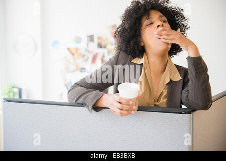 Businesswoman with cup of coffee yawning in cubicle Stock Photo