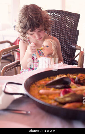 Girl sitting at dinner table pretending to feed her doll Stock Photo