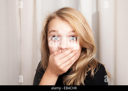 Beautiful blond Caucasian surprised girl opened her eyes wide and covers her mouth with her hands, closeup studio portrait Stock Photo