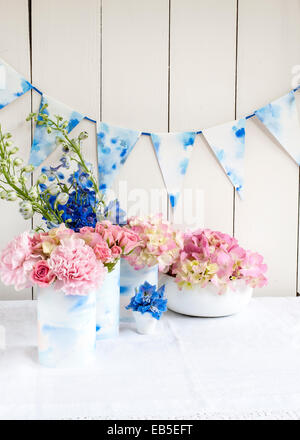 blue delphinium, pink roses, hydrangeas, carnation in tins decorated with painted watercolour paper, paper bunting, fabric Stock Photo