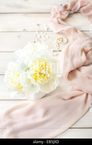 A bunch of white creamy peonies in a white jug on a white wooden table with a light pink scarf and pearls in the background Stock Photo