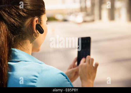 Mid adult hispanic person with mobile phone and bluetooth headset, typing on telephone in the street Stock Photo