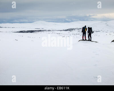 two men with heavy packs wearing snowshoes standing looking out over a  winter mountain landscape Stock Photo