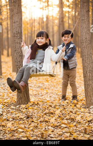 Little boy pushing his sister on a swing in autumn woods Stock Photo