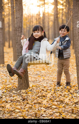 Little boy pushing his sister on a swing in autumn woods Stock Photo