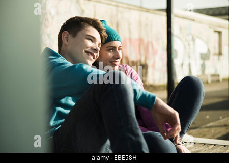 Teenage girl and boy sitting on ground, smiling and looking at view together, Mannheim, Germany Stock Photo