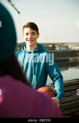 Teeange boy holding basketball outdoors, smiling and looking at teenage girl, industrail area, Mannheim, Germany Stock Photo