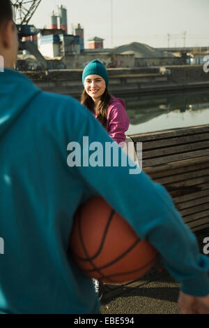 Teenage girl outdoors wearing toque, smiling and looking at teenage boy holding basketball, industrial area, Mannheim, Germany Stock Photo