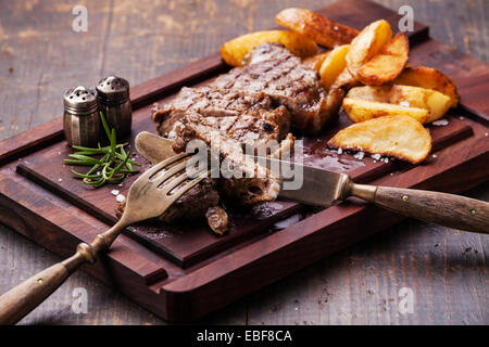 Sliced well done grilled New York steak with roasted potato wedges on cutting board on dark wooden background Stock Photo