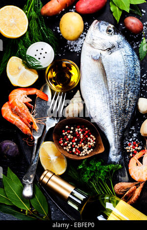 Delicious fresh fish and seafood on dark vintage background. Fish, cockles and shrimps with aromatic herbs, spices & vegetables Stock Photo