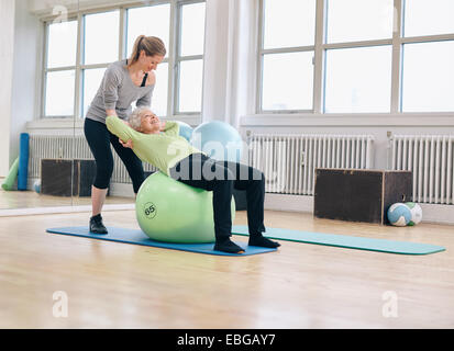 Female coach helping senior woman exercising in gym. Elder woman working out on pilates ball at health club being assisted by he Stock Photo