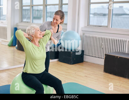 Mature woman working out on a fitness ball with help from personal trainer at gym. Happy senior woman exercising with instructor Stock Photo