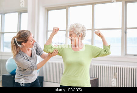 Portrait of female trainer looking at elderly woman flexing her bicep at the rehabilitation center. Happy about her recovery. Stock Photo