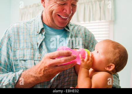 Grandfather bottle feeding baby granddaughter on lap in living room Stock Photo