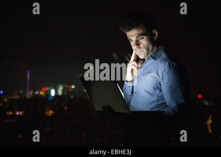 Young businessman looking at laptop in front of skyscraper office window at night, Shanghai, China Stock Photo