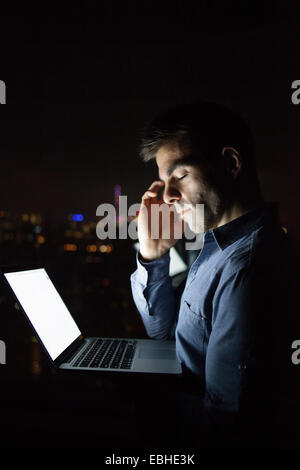 Tired young businessman with laptop in front of skyscraper office window at night, Shanghai, China Stock Photo