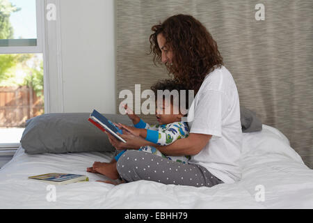 Young woman sitting on bed reading book to toddler son Stock Photo