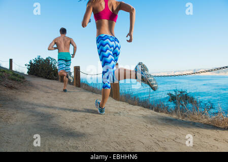 Mid adult man and young woman running on path by sea, rear view