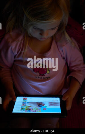 Blond child girl using (playing, watching) digital tablet on couch at night Stock Photo