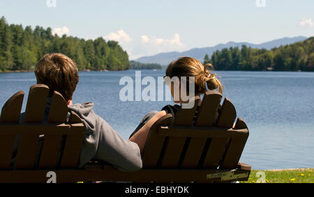 Young couple man woman sitting in double Adirondack chair over looking Long Lake New York USA. Adirondack State Park Adirondacks Stock Photo