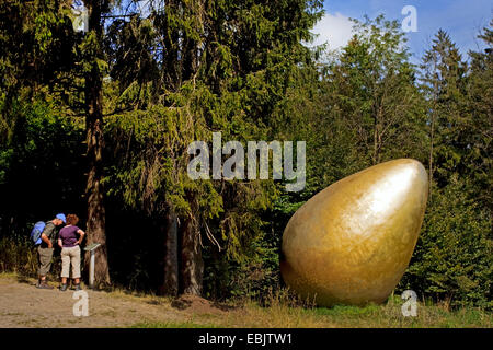 two wanderers on the Waldskulpturenweg reading the information plate of the sculpture 'Was war zuerst?' ('What came first?') which shows a giant golden egg, Germany, North Rhine-Westphalia, Siegerland, Bad Berleburg Stock Photo