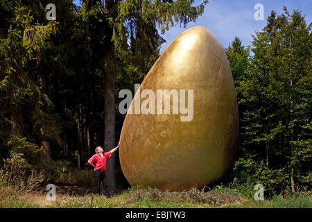 man on the Waldskulpturenweg leaning against the sculpture 'Was war zuerst?' ('What came first?') which shows a giant golden egg, Germany, North Rhine-Westphalia, Siegerland, Bad Berleburg Stock Photo