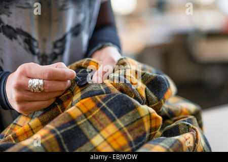 Close up seamstress hand sewing button onto tartan jacket in workshop Stock Photo