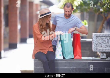 Young woman sitting on wall with shopping bags, man looking in bags Stock Photo