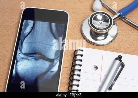 Technology use in healthcare. Smartphone displays an X-ray CT scan of a knee, acoustic stethoscope, notepad, and pen on doctors office desk Stock Photo