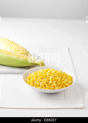 Raw corncobs on marble cutting board and bowl of boiled sweetcorn Stock Photo