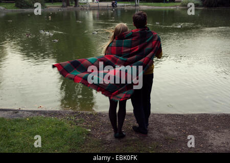 Rear view of young couple wrapped in blanket on park lakeside Stock Photo