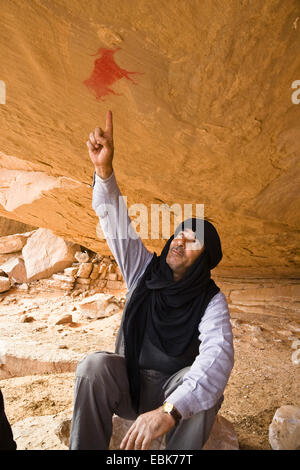Touareg sitting under a rock spur pointing at prehistoric rock paintings at the Acacus Mountains, Libya, Sahara Stock Photo