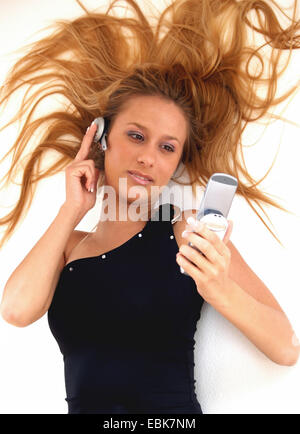 young woman lying on the ground with flowing hair while holding an earphone against her ear and looking at the mobile in her hand Stock Photo