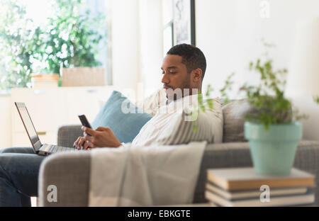 Man sitting in living room, using laptop and cell phone Stock Photo