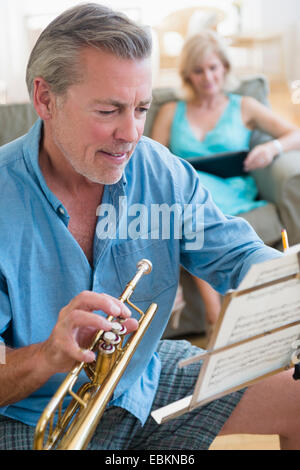 Portrait of senior man holding trumpet looking at music stand with woman on sofa in background Stock Photo