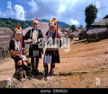 Three Akha girls in a mountain village, in traditional costume and headdress, bamboo huts with thatched roofs at back Stock Photo