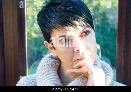Beautiful young woman with short, black hair looking out of a window deep in thought with garden in the background and patio door Stock Photo