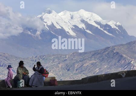 Aymara women waiting for public bus on the route to El Alto with Illimani peak in background, Bolivia Stock Photo