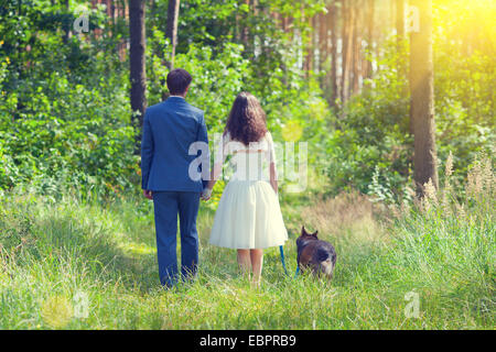 Young newlyweds holding hands walking with dog in the forest back to camera Stock Photo