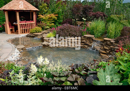 The pond area in a reflective aquatic garden with planted rockery, waterfalls and a wooden Summer House Stock Photo