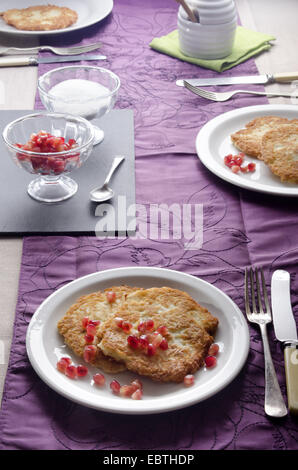 laid table with rustic home made potato pancakes, sugar in a bowl on slate, fresh pomegranate seed, lilac table runner, fork, kn Stock Photo