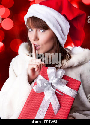 Young woman in Santa's cap keeps Christmas present wrapped in red paper on black background. She making keep quiet gesture Stock Photo
