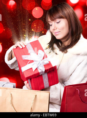 Young joyful woman opens Christmas present wrapped in red paper on red blurred background. From gift box goes magic light Stock Photo