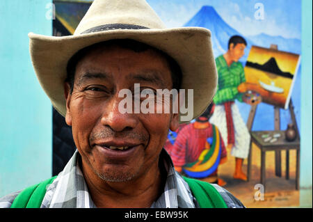 man is standing in front of a painting on a house wall showing a man painting a picture and a woman weaving a carpet, Guatemala, Atitlan lake, Santa Cruz la Laguna Stock Photo